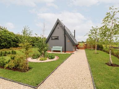 A gravel pathway leading to a small gray triangular house with a bench and surrounding trees and bushes at Silver Birch Lodge in Misterton