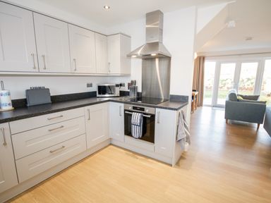 A kitchen with white cabinets black countertops an oven microwave a knife block and a living room with gray sofas at 14 Parc Delfryn in Brynteg near Benllech