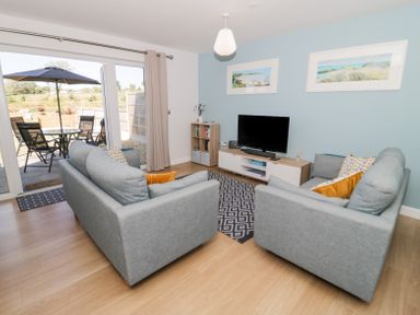 A living room with two gray sofas a TV on a stand a small bookcase and sliding doors leading to a patio with a table and chairs at 14 Parc Delfryn in Brynteg near Benllech