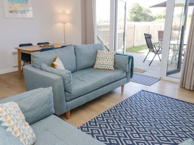 A sitting area with two blue sofas and patterned pillows a wooden dining table with chairs a floor lamp and glass doors opening to a patio with outdoor chairs at 14 Parc Delfryn in Brynteg near Benllech