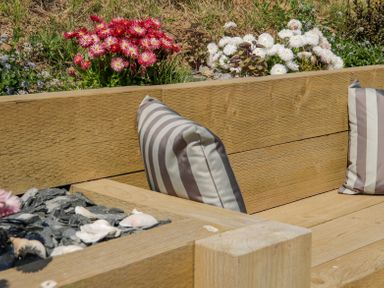 An outdoor wooden bench with striped cushions near flower beds with red and white flowers at 14 Parc Delfryn Brynteg near Benllech