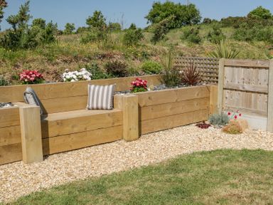 A wooden bench with striped cushions on a gravel area next to a garden bed with flowers and shrubs at 14 Parc Delfryn in Brynteg near Benllech