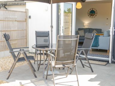 A patio with a round glass table and four chairs next to an open door showing a living room with sofas and wall clock at 14 Parc Delfryn in Brynteg near Benllech
