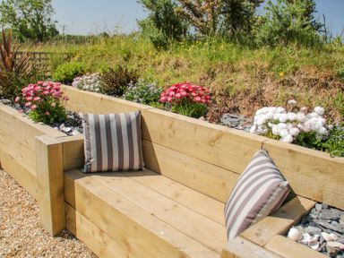 A wooden outdoor bench with striped cushions and flower beds behind it at 14 Parc Delfryn in Brynteg near Benllech