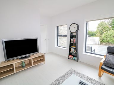 A living room with a television on a wooden stand a clock bookshelf chair and large windows at Wee Andy's in Slavary near Buncrana County Donegal