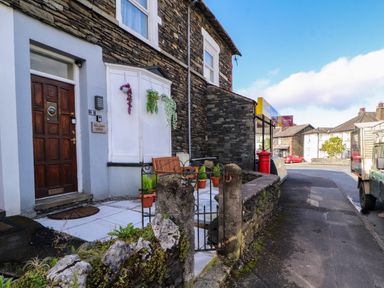 An exterior view of a stone house with a wooden door a bench and hanging plants near a street at Neland View in Windermere
