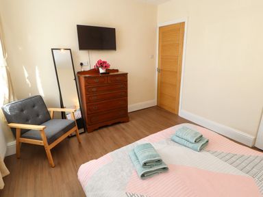 A bedroom with a bed with folded towels a wooden chair a standing mirror a wooden chest of drawers with flowers and a wall mounted television at Neland View in Windermere