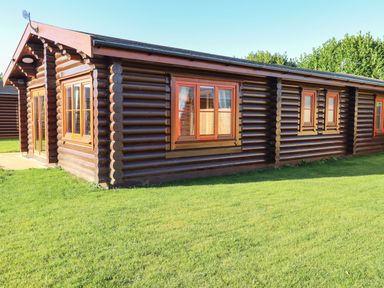 An exterior view of a wooden log cabin with multiple windows and a grass lawn at Elm Lodge Greetham Rutland