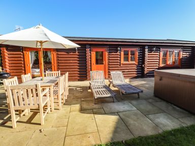 A patio area with wooden dining table and chairs under umbrella two wooden loungers and a hot tub outside a log cabin at Elm Lodge in Greetham Rutland