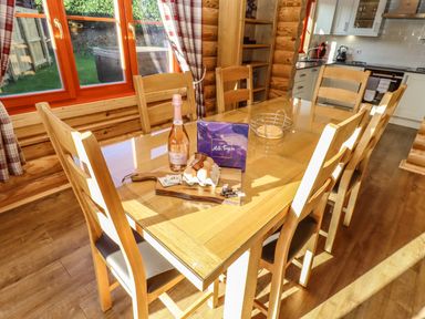 A dining area with a wooden table and six chairs with items including a bottle of prosecco and a box of milk tray chocolates at Elm Lodge in Greetham Rutland