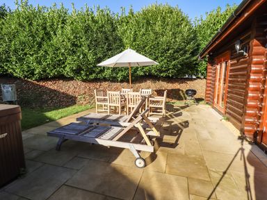An outdoor patio with wooden lounge chairs a dining table with umbrella a barbecue and a wooden log cabin at Elm Lodge in Greetham Rutland