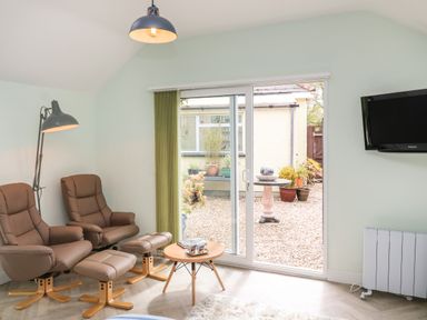 A small sitting area with two brown armchairs and matching footstools a small table a floor lamp a wall-mounted TV and sliding glass doors leading to a garden at Byghan Thornton in Illogan