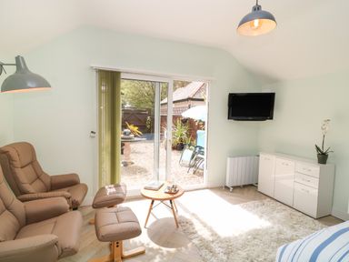 A room with beige armchairs a small round table a white cabinet a wall-mounted TV and glass sliding doors leading outside at Byghan Thornton in Illogan