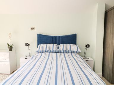 A bedroom with a bed that has a blue headboard and striped blue and white bedding two bedside tables with lamps and a white dresser with a plant at Byghan Thornton in Illogan