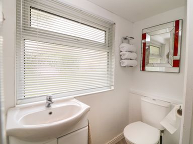 A bathroom with a sink under a window with blinds a toilet a mirror and rolled towels on a rack at Byghan Thornton in Illogan