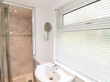 A bathroom with a white sink faucet a shower with glass doors and tiled walls and a window with white blinds at Byghan Thornton Illogan