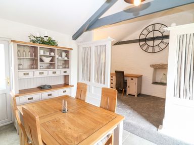 A dining area with a wooden table and chairs and a cabinet with glassware next to a carpeted study space with a desk chair and large wall clock at Polgear View Polgear Nr Redruth