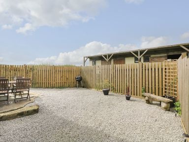 A fenced outdoor area with gravel ground a wooden table and chairs a charcoal grill two potted plants and a stone bench at Polgear View in Polgear Nr Redruth