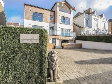 A modern three story building with balconies and a paved parking area with a lion statue and a sign saying 1-3 Min Manton at Bryn Min Manton Apartment in Caernarfon