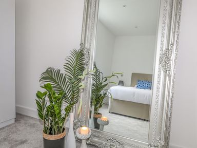A bedroom with a bed with a blue pillow reflected in a large ornate mirror next to potted plants and a lit candle at Bryn Min Manton Apartment in Caernarfon