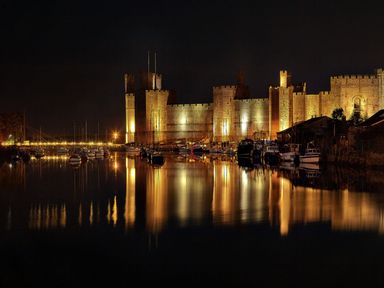 A nighttime view of a castle with boats docked in front and their reflections at Bryn Min Manton Apartment in Caernarfon