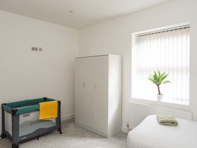 A simple bedroom with a wardrobe a crib a bed and a plant on the window sill at Bryn Min Manton Apartment in Caernarfon