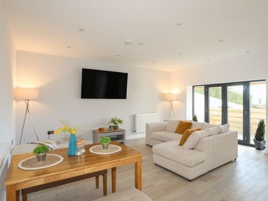 A living room with a beige sofa and yellow pillows next to glass doors and a wooden dining table with plants at Bryn Min Manton Bach in Caernarfon