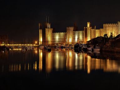 A nighttime view of a castle illuminated with lights next to a harbor with boats and their reflections in the water at Bryn Min Manton Bach in Caernarfon