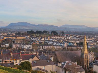 A town with rows of houses and a church with a steeple with hills in the background at Bryn Min Manton Bach in Caernarfon