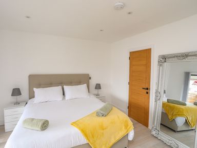 A bedroom with a bed with white sheets and pillows bedside tables with lamps a yellow blanket and a green towel a wooden door and a large ornate silver mirror at Bryn Min Manton Bach in Caernarfon