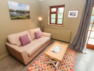 A living room with a beige sofa with purple cushions a wooden coffee table a patterned rug a floor lamp two framed pictures on the wall and windows at Bwthyn Canol in Haverfordwest