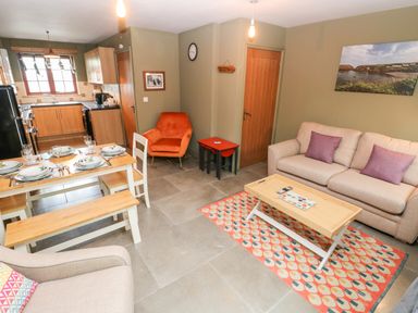 An open plan living room and kitchen with a dining table set for six an orange armchair a beige sofa with purple cushions a wooden coffee table on a patterned rug and kitchen cabinets with appliances at Bwthyn Canol in Haverfordwest