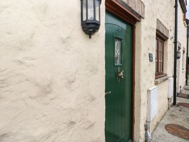 A close-up of a green door with a decorative knocker set in a cream textured wall with a window and outdoor light at Bwthyn Canol in Haverfordwest