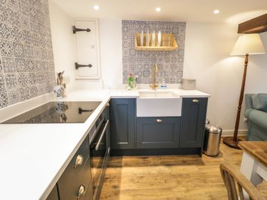 A kitchen with blue cabinets a white countertop a sink a stovetop a wooden shelf with plates and glasses and a floor lamp next to a sofa at Gelli Bach at Gelli Las in Llanbedr