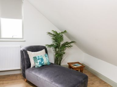 A chaise lounge with pillows beside a window and a plant with a small wooden table under a slanted ceiling at Ysgoldy Brynwyre in Llanrhystud