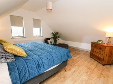 A bedroom with a bed a blue quilt two windows a wooden chest of drawers with a lamp and a plant a chair and wooden floor at Ysgoldy Brynwyre in Llanrhystud