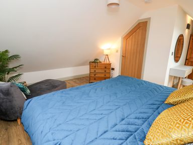 A bedroom with a blue bedspread yellow pillows a wooden chest of drawers a lamp a gray chair a plant and a wooden door at Ysgoldy Brynwyre in Llanrhystud