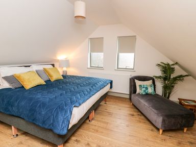 A bedroom with a large bed with blue quilt and yellow pillows and a chaise lounge next to two windows at Ysgoldy Brynwyre in Llanrhystud
