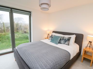 A bedroom with a bed with cushions and a blanket two bedside tables with lamps and a large window showing grass at Ysgoldy Brynwyre in Llanrhystud