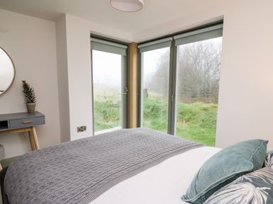 A bedroom with a bed gray blanket green pillows next to large windows and a small table with a plant at Ysgoldy Brynwyre in Llanrhystud