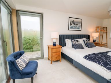 A bedroom with a blue upholstered bed and pillows two wooden nightstands with lamps a blue armchair and glass door showing grass outside at Ysgoldy Brynwyre in Llanrhystud