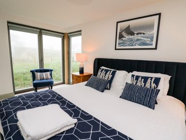 A bedroom with a bed with blue patterned pillows and a blanket a blue chair and large windows at Ysgoldy Brynwyre in Llanrhystud