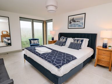 A bedroom with a bed with blue and white bedding two wooden nightstands with lamps a blue chair and large windows at Ysgoldy Brynwyre in Llanrhystud