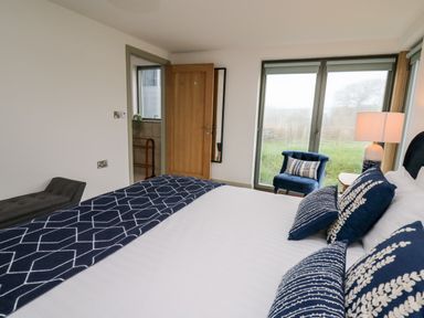 A bedroom with a bed with blue patterned pillows and throw a blue chair near two glass doors with a lamp on a small table at Ysgoldy Brynwyre in Llanrhystud