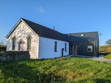 A white stone building with three arched windows and a purple door attached to a modern dark gray building at Ysgoldy Brynwyre in Llanrhystud