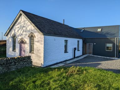 A building with white stone walls and purple door next to a modern black metal extension surrounded by grass at Ysgoldy Brynwyre in Llanrhystud