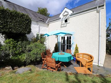 An outdoor patio area with a round table covered by a teal tablecloth and an umbrella surrounded by wooden chairs next to a white house at Tyddyn Bach Newport Pembrokeshire