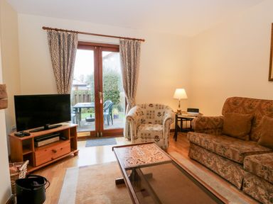 A living room with a TV on a wooden stand a patterned armchair a brown sofa a glass coffee table and a lamp by the sofa at Friars Fold in Newby Bridge