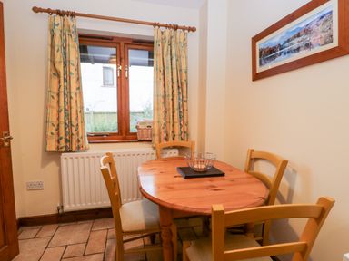 A dining area with a wooden table and four chairs near a window with yellow patterned curtains at Friars Fold in Newby Bridge
