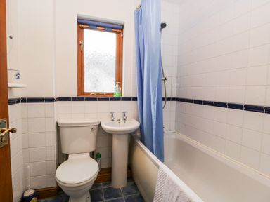 A bathroom with a toilet, a pedestal sink, a bathtub with a blue shower curtain, a towel hanging on the bathtub, and a window with a roller blind at Friars Fold in Newby Bridge